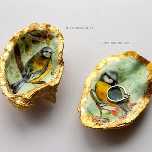 May include: Two gold-leafed oyster shell ring dishes with bluebird designs. One dish has a ring inside. The text "BIRD DESIGN 01" and "BIRD DESIGN 02" is above the dishes.