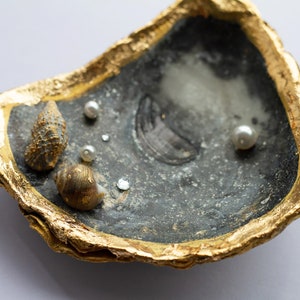 May include: A black and gold oyster shell dish with a gold rim. The dish is decorated with small white pearls and seashells.