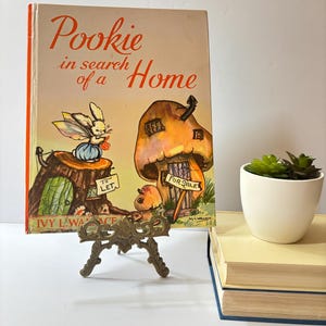 May include: A vintage children's book, "Pookie in Search of a Home", featuring a fairy bunny and a mushroom house illustration. The book is displayed on a decorative brass stand. A small potted plant and books are in the background.