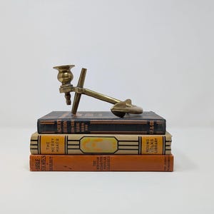 May include: A brass anchor candle holder with a single candle cup. The holder is sitting on top of a stack of three vintage books. The books are titled "The Bible Stories", "The Merry Maker", and "Sun-Gray Basic Book One".