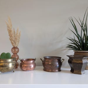 Peut inclure: Quatre pots en métal différents, deux en cuivre, un en laiton et un en métal foncé. Les pots sont disposés sur une surface blanche. Un bouquet d'herbe sèche dans un vase en verre rose est à gauche des pots. Une plante serpent en pot est à droite des pots.