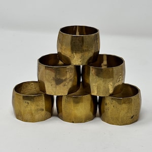 May include: Six brass napkin rings stacked. Each ring is cylindrical with a faceted exterior, giving them a geometric look. The rings have a warm, golden colour and are arranged in a pyramid shape.