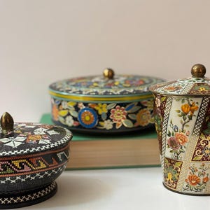 May include: Three antique decorative tins with lids. The tins feature floral and geometric patterns in various colours, including black, gold, and orange. The tins are of different shapes and sizes, with a round, a cylindrical, and a bowl-shaped tin.