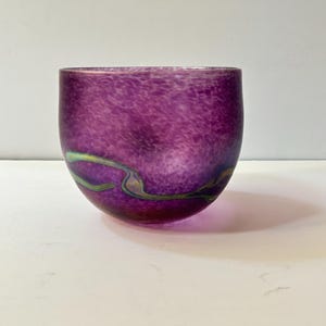 May include: A hand-blown glass bowl in shades of purple with a speckled pattern. The bowl has a rounded shape with a wide opening. Green and gold swirls accent the lower portion of the bowl. The bowl is sitting on a white surface.