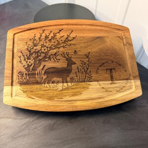Könnte beinhalten: Holzschneidebrett mit Hirsch- und Baumdesign. Das Brett hat ein geschnitztes Bild eines Hirsches unter einem Baum, mit dem Buchstaben "T" auf der rechten Seite. Rechteckig mit abgerundeten Ecken und einer Saftrille.