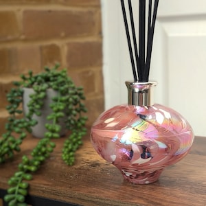 Könnte beinhalten: Rosa Glas-Diffuserflasche mit silbernem Verschluss und schwarzen Rattanstäben. Die Flasche hat ein Spiraldesign und eine irisierende Oberfläche.