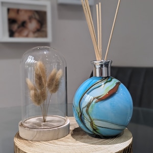 Può includere: Un diffusore di profumo in vetro blu con un coperchio in legno e una cupola di vetro contenente erba secca. La bottiglia del diffusore ha un motivo a spirale di blu, verde e bianco. La bottiglia del diffusore è appoggiata su una base di legno.
