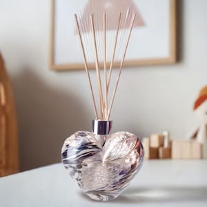 Op de afbeelding: Een hartvormige glazen diffuser met een zilveren deksel en houten stokjes. De diffuser heeft een zwart-wit marmerpatroon met een vleugje roze.