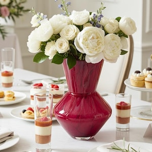 May include: An elegant table setting featuring a burgundy vase with white roses. Small desserts and layered parfaits are presented on plates and in glasses. Silverware and folded napkins create a refined atmosphere.