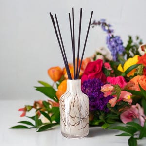 Może przedstawiać: Biała butelka dyfuzora z czarnymi trzcinami i marmurowym wzorem. Butelka jest otoczona bukietem kolorowych kwiatów, w tym różowych, fioletowych, żółtych i pomarańczowych.