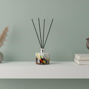 Op de afbeelding: Een witte plank toont een kleurrijke glazen diffuser met zwarte rietjes, een kleine vaas met gedroogd gras, een bronzen vogelbeeldje en twee boeken. De diffuser heeft een zilveren bovenkant en een abstract ontwerp.