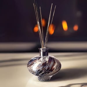 Puede incluir: Un difusor decorativo de vidrio con varillas plateadas. El recipiente de vidrio estampado presenta remolinos de plata, marrón y morado. El difusor se encuentra sobre una superficie blanca, con un fondo borroso de una chimenea.