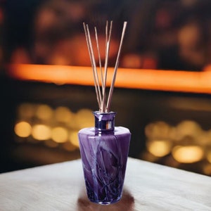 Può includere: Bottiglia diffusore in vetro viola con un motivo a spirale e bastoncini di legno. La bottiglia è appoggiata su un tavolo di legno.