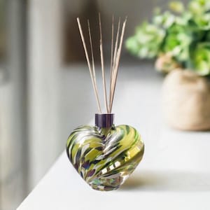 Può includere: Un diffusore in vetro a forma di cuore con un design a spirale verde e oro. Il diffusore ha un collo viola scuro e contiene diversi bastoncini marroni. Lo sfondo include una vista sfocata di verde in un vaso beige.