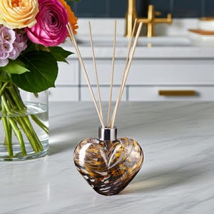 May include: A heart-shaped glass diffuser with a silver collar, filled with reeds. The glass has a brown and gold marbled pattern. A bouquet of colourful flowers in a glass vase is in the background.