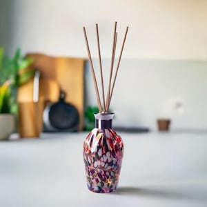 Könnte beinhalten: Eine dekorative Glas-Diffusorflasche mit einem bunten, gesprenkelten Design in Rot-, Lila- und Grüntönen. Die Flasche hat einen silbernen Kragen und enthält mehrere Holz-Diffusorstäbchen. Das Produkt ist für die Aromatherapie.