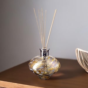 Può includere: Una bottiglia di diffusore in vetro con un tappo argentato e un motivo a spirale in tonalità oro, bianco e marrone. La bottiglia è riempita con un liquido trasparente e ha bastoncini di legno inseriti.