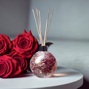 May include: A red glass diffuser bottle with a marbled pattern and wooden sticks. The bottle is surrounded by red roses.