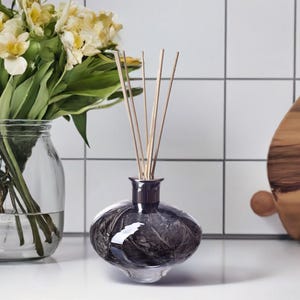 Op de afbeelding: Een zwarte en zilveren glazen diffuserfles met een rond, getextureerd ontwerp. De fles is gevuld met een heldere vloeistof en heeft houten stokjes erin gestoken. De diffuser staat op een wit aanrecht.