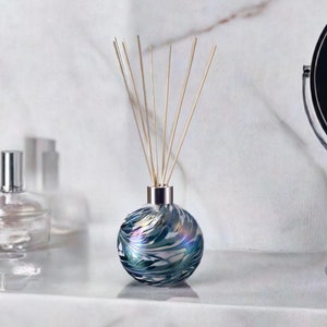 May include: A spherical glass diffuser with a silver collar and wooden reeds. The iridescent glass bottle has swirling patterns of blue, green, and silver. Other glass bottles are in the background.