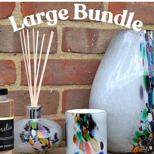 Może przedstawiać: Duży zestaw produktów zapachowych do domu. Zestaw zawiera dyfuzor z srebrną nakrętką i szklaną butelką o kolorowym wzorze, świecę z białym wierzchołkiem i kolorowym szklanym słoikiem oraz przezroczystą szklaną butelkę z etykietą "Amelia Creamy Vanilla".