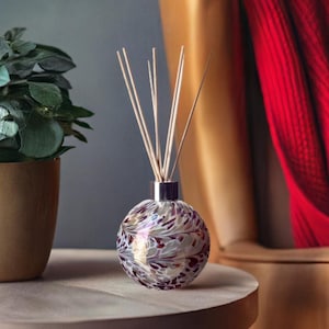 Op de afbeelding: Een ronde glazen diffuserfles met een wit en gouden swirl-patroon en rode accenten. De fles is gevuld met een heldere vloeistof en heeft houten stokjes die uit de bovenkant steken.