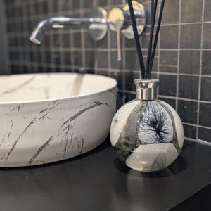 Könnte beinhalten: Eine schwarze und weiße Marmor-Diffuserflasche mit schwarzen Rattanstäben. Die Flasche hat einen silbernen Verschluss und einen klaren Glasboden. Die Flasche steht auf einer schwarzen Oberfläche.
