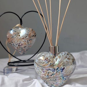 Può includere: Un diffusore in vetro a forma di cuore con un colletto argentato e bastoncini di legno. Il vetro presenta un motivo a spirale di colori beige, bianco e iridescenti. Un ornamento a forma di cuore è appeso sullo sfondo, anch'esso con un motivo a spirale.