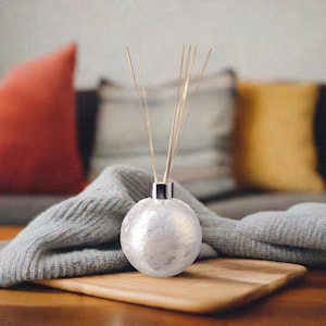 Può includere: Un flacone diffusore bianco di forma rotonda con un tappo argentato. Il flacone è riempito con un liquido trasparente e ha un set di bastoncini di legno inseriti al suo interno. Il flacone è appoggiato su una superficie di legno.