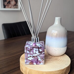 Puede incluir: Una botella difusora cuadrada de vidrio con un dise&ntilde;o de espiral morado y blanco. La botella est&aacute; llena de un l&iacute;quido transparente y tiene varillas difusoras grises. La botella est&aacute; sentada sobre una base de madera.