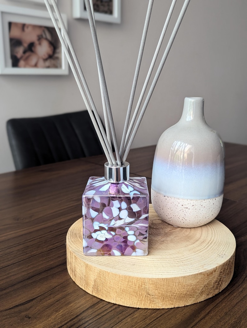 Puede incluir: Una botella difusora cuadrada de vidrio con un dise&ntilde;o de espiral morado y blanco. La botella est&aacute; llena de un l&iacute;quido transparente y tiene varillas difusoras grises. La botella est&aacute; sentada sobre una base de madera.
