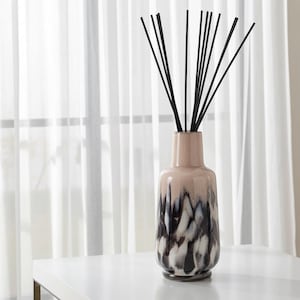 May include: A decorative vase with black reeds. The vase is a light pink colour at the top, transitioning to a pattern of black, white, and grey. The vase is on a white surface in front of a white curtain.