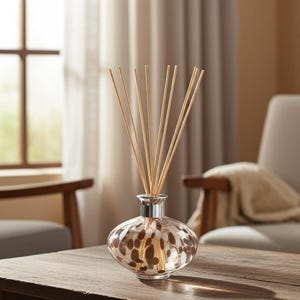 Könnte beinhalten: Ein transparenter Glas-Raumduft-Diffusor mit einem abgerundeten, flachen Boden, der ein internes Muster aus braunen und weißen ovalen Flecken aufweist. Die Flasche hat einen silberfarbenen Metallkragen und enthält mehrere dünne, helle Holzstäbchen.