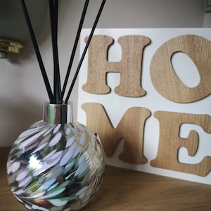 Puede incluir: Un difusor decorativo de vidrio con varillas negras, cuello plateado y diseño moteado colorido. El difusor está sobre una estantería de madera junto a un letrero blanco con la palabra "HOME" en letras de madera. Decoración del hogar.