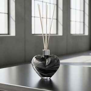 Puede incluir: Un difusor de vidrio veteado en blanco y negro con forma de corazón, con un collar plateado, que contiene varias cañas de color claro. El difusor se encuentra sobre una superficie oscura, con un fondo borroso de ventanas y una pared de hormigón.