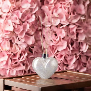 Op de afbeelding: Een hartvormige, parelmoerwitte diffuserfles met een zilveren kraag, gevuld met riet. De fles staat op een houten oppervlak, met een achtergrond van roze bloemen. De diffuser is een decoratief geurartikel voor in huis.