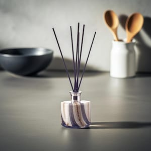 May include: A decorative reed diffuser with dark purple reeds. The glass bottle has a striped pattern in white, navy, and tan. A black bowl and a white jar with wooden spoons are in the background.