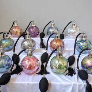 Puede incluir: Una colección de frascos de perfume decorativos de vidrio con tapas plateadas y atomizadores de bulbo negros. Los frascos redondos presentan colores iridiscentes, incluyendo rosa, azul, verde y dorado. Los frascos están dispuestos sobre una superficie blanca.