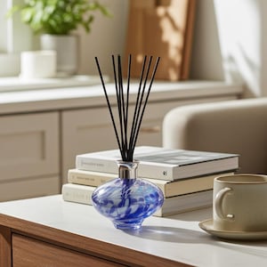 May include: A blue and white glass diffuser bottle with black reeds. The bottle has a silver neck and sits on a white surface next to a stack of books and a beige mug. The diffuser is a decorative home fragrance item.