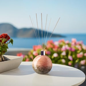 Op de afbeelding: Een ronde glazen diffuserfles in roze en bruin met zilveren rietjes. De fles heeft een getextureerd oppervlak. De fles staat op een witte tafel.