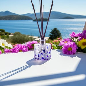 May include: A glass reed diffuser bottle with a speckled blue and white pattern, featuring a silver-toned collar and several thin dark brown reeds extending upward, surrounded by vibrant pink, yellow, and white flowers on a white surface.