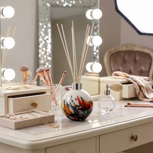 Può includere: Un tavolo da trucco con un diffusore decorativo, pennelli per il trucco e un flacone di profumo. Il diffusore ha un design bianco e colorato. Uno specchio con luci è sullo sfondo. Un vassoio portagioie con collane e orecchini è sul tavolo.