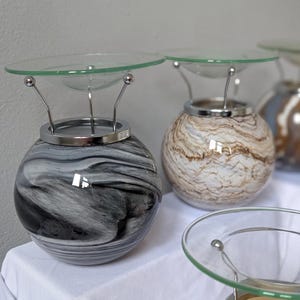 May include: Decorative glass oil warmers with a round, marbled base in shades of black and white, and brown and white. Each warmer has a clear glass dish supported by silver metal arms and a silver metal rim.