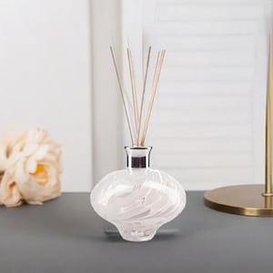 May include: A white glass diffuser bottle with a silver neck and tan reeds. The bottle has a distinctive, rounded shape and a marbled white design. The diffuser is placed on a grey surface.