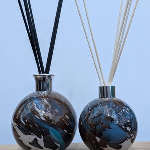 Op de afbeelding: Twee decoratieve glazen diffuserflessen met zilverkleurige doppen. Eén fles heeft zwarte stokjes, de andere heeft naturelkleurige stokjes. De flessen hebben een gemarmerd ontwerp in bruin, blauw en wit.