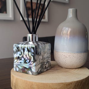 Könnte beinhalten: Eine quadratische Glasdiffusor-Flasche mit silbernem Deckel und schwarzen Schilfrohren. Die Flasche hat ein wirbelndes Muster in Schwarz, Weiß und irisierenden Farben. Der Diffusor steht auf einem Holzsockel neben einer grauen Keramikvase.