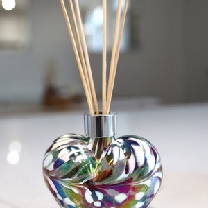 Puede incluir: Un difusor de aroma de vidrio en forma de coraz&oacute;n con una tapa plateada y palitos difusores de madera. La botella es iridiscente y presenta un patr&oacute;n de remolino de blanco, verde y morado.