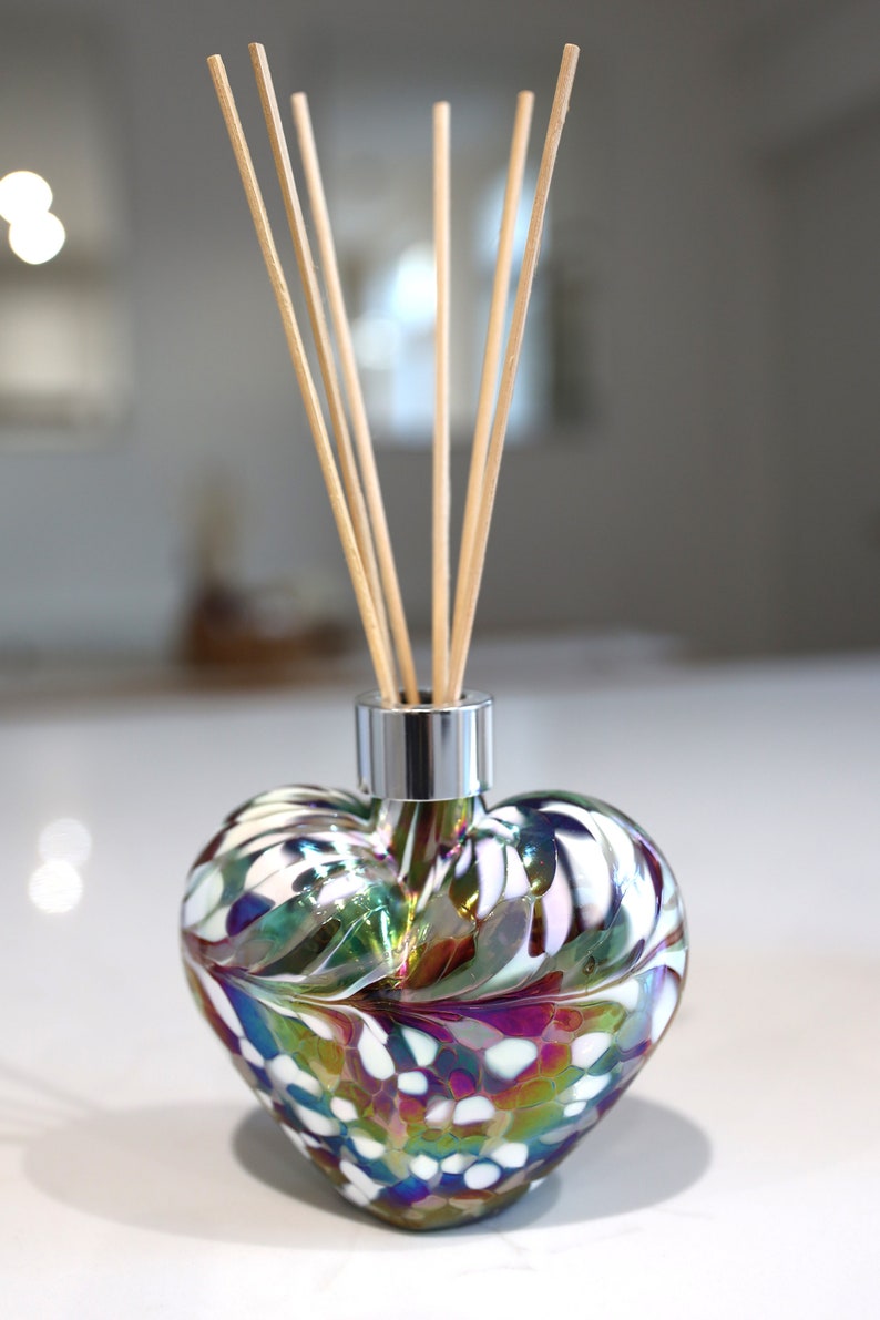 Puede incluir: Un difusor de aroma de vidrio en forma de coraz&oacute;n con una tapa plateada y palitos difusores de madera. La botella es iridiscente y presenta un patr&oacute;n de remolino de blanco, verde y morado.