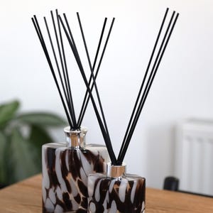 Può includere: Due diffusori a bastoncino decorativi con bastoncini neri. Le bottiglie di vetro hanno una forma quadrata e un motivo maculato marrone e bianco. I diffusori poggiano su una superficie di legno, con sfondo bianco.