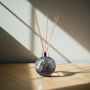 Può includere: Un diffusore decorativo con un flacone rotondo in vetro con motivo marmorizzato nei toni del verde, marrone e blu. Il flacone ha un colletto color oro rosa ed è riempito di bastoncini di legno.